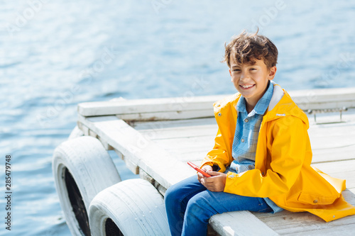 Cheerful dark-eyed boy feeling happy while sitting near river