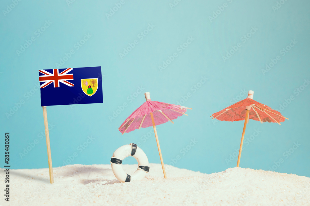 Miniature flag of Turks And Caicos Islands on beach with colorful umbrellas and life preserver. Travel concept, summer theme.