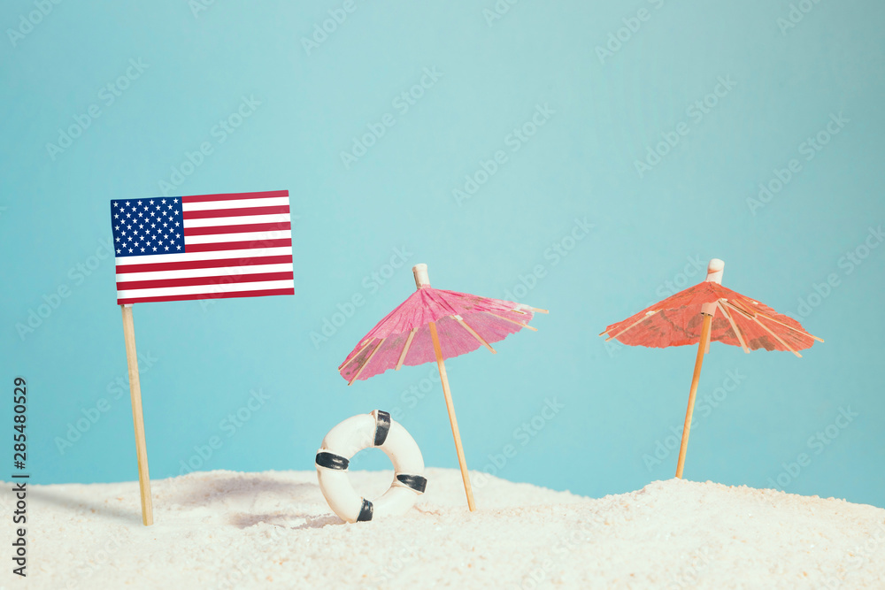 Miniature flag of United States on beach with colorful umbrellas and life preserver. Travel concept, summer theme.