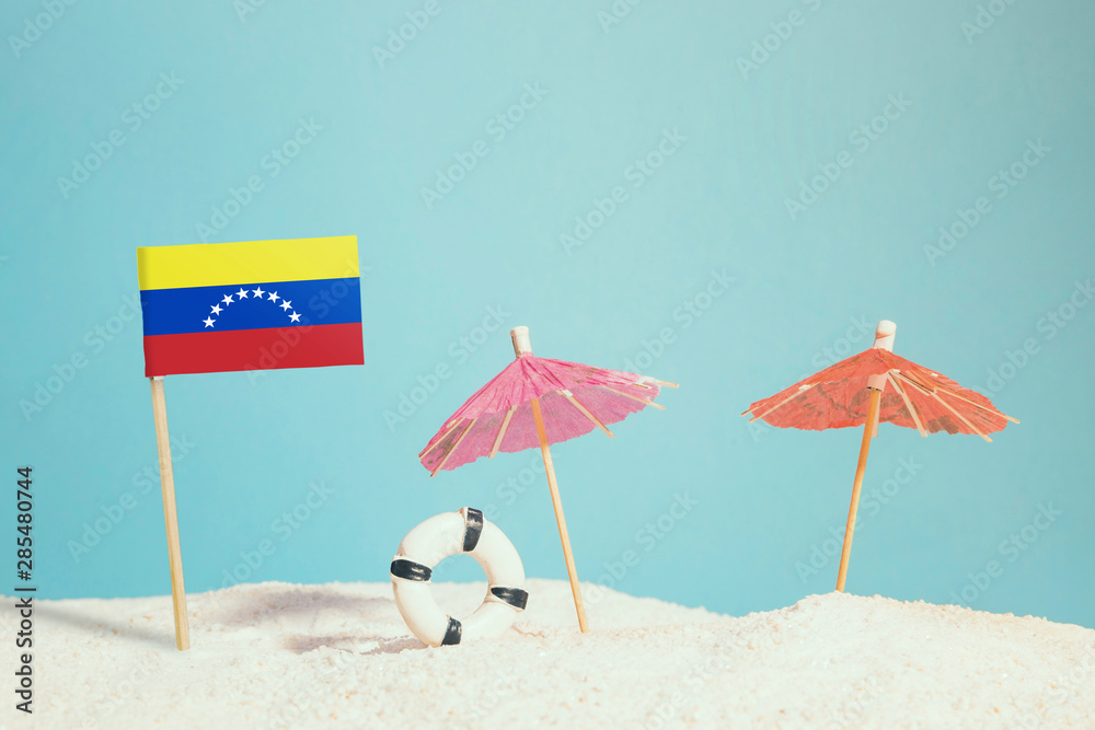 Miniature flag of Venezuela on beach with colorful umbrellas and life preserver. Travel concept, summer theme.