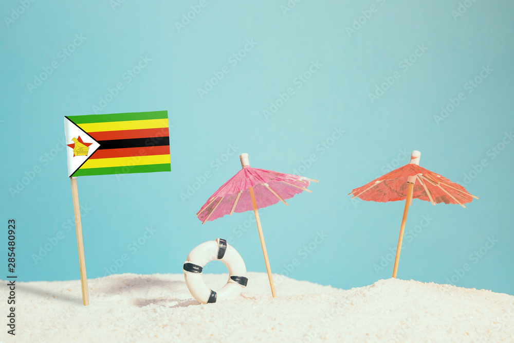 Miniature flag of Zimbabwe on beach with colorful umbrellas and life preserver. Travel concept, summer theme.