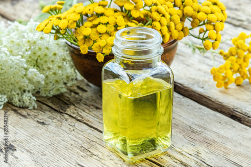 Glass bottle of essential oil or tincture of extract near yellow fresh flowers of tansy and yarrow on wooden background. Herbal medicine concept.