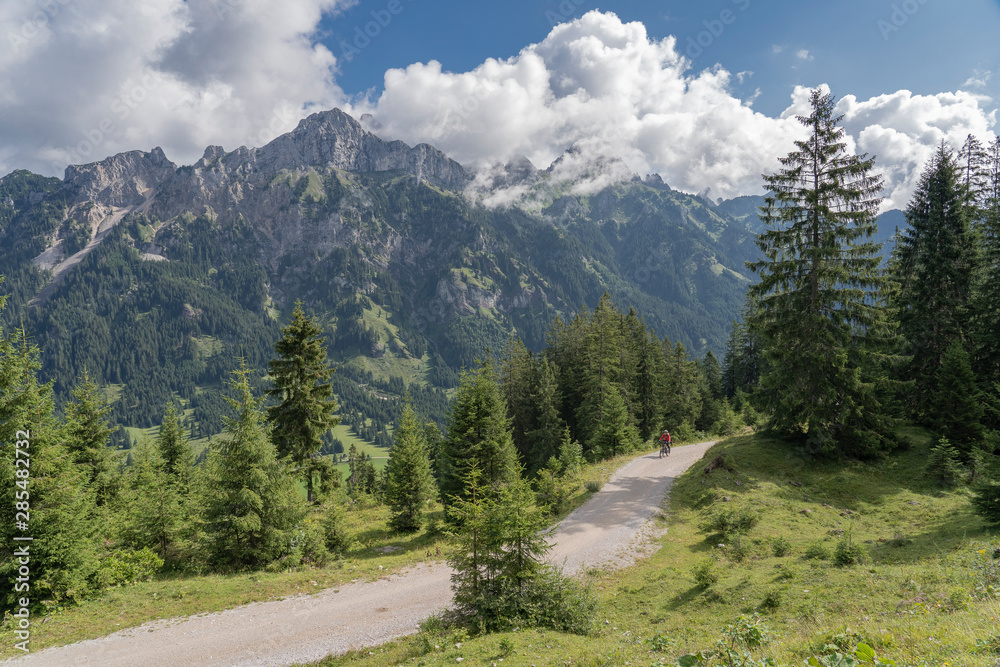 nice and active senior woman, riding her e-mountain bike in the Tannheim valley , Tirol, Austria, with the village of Tannheim and famous summits Gimpel and Rote Flueh