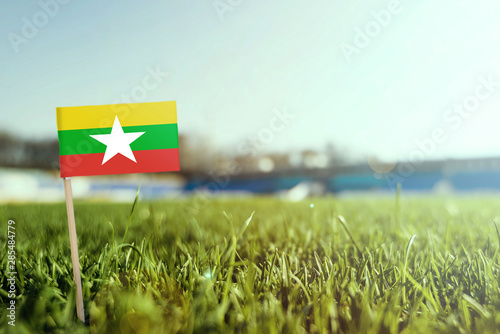 Miniature stick Myanmar flag on green grass, close up sunny field. Stadium background, copy space for text.