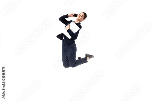 smiling businesswoman dancing while talking on smartphone and holding laptop isolated on white