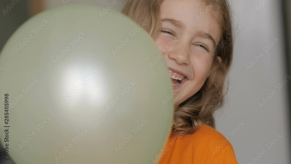 Little girl looks out from behind balloon and laughs happily. Close up portrait of a happy little child bursting in laugh. Child girl blonde with blue eyes and laughs indoors. Closeup. Slow Motion. Little girl looks out from behind balloon and laughs happily. Close up portrait of a happy little child bursting in laugh. Child girl blonde with blue eyes and laughs indoors. Closeup. Slow Motion.