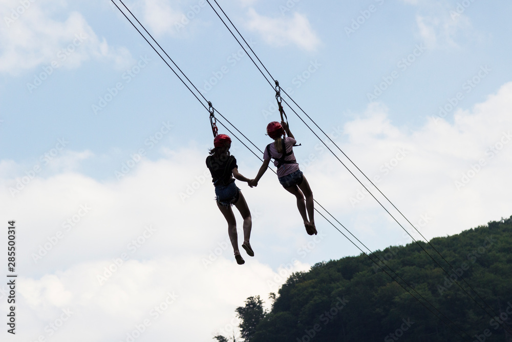 Two girls hold hands and walk along a zip line, a sport in an extreme park. Two girls hold hands and walk along a zip line, a sport in an extreme park.