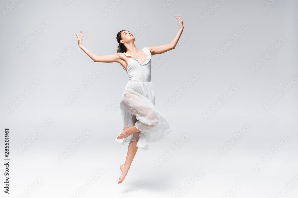 young elegant ballerina in white dress dancing on grey background