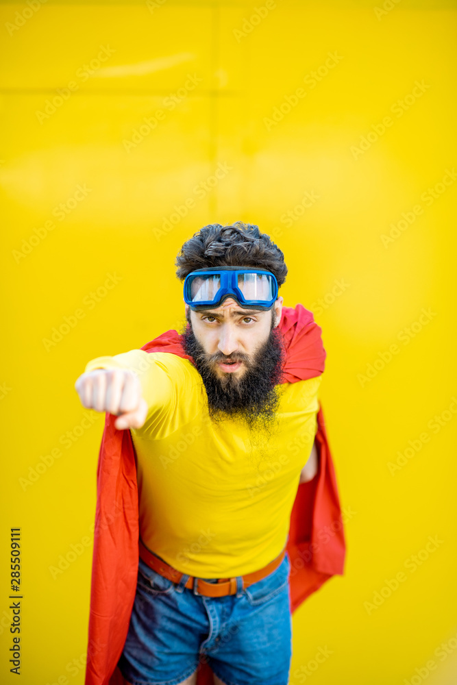 Fototapeta premium Portrait of a man like a superhero in colorful clothes and pilot's glasses on the yellow background