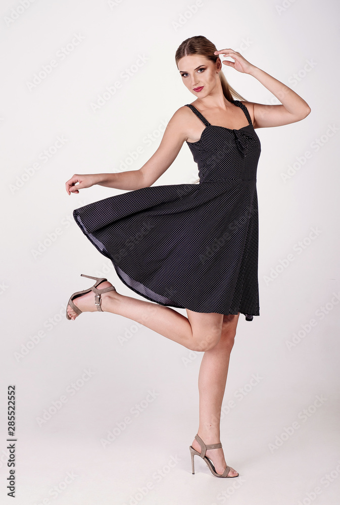 Naklejka premium Young attractive sexy blond woman model with light hair, wearing short black dress and sandals on heels, posing and dancing in white studio