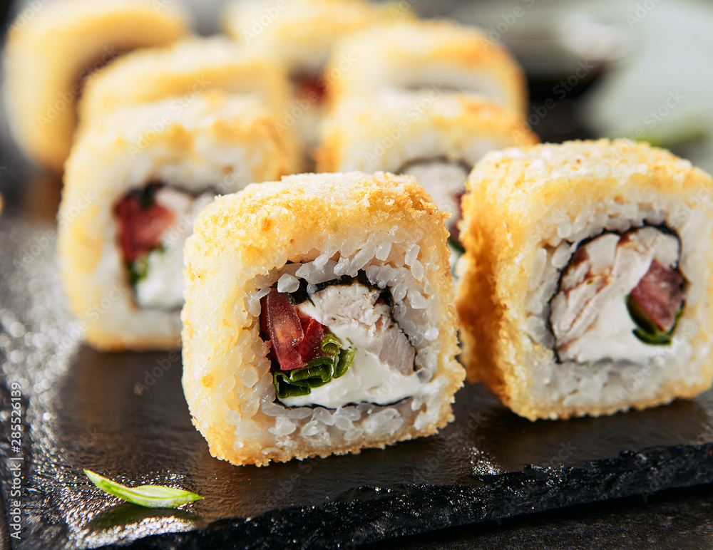 Macro shot of tempura uramaki sushi rolls with smoked chicken, cream cheese, green onion, tomato, rice and nori. Chopsticks holding hot crispy maki roll on black stone slate background closeup Macro shot of tempura uramaki sushi rolls with smoked chicken, cream cheese, green onion, tomato, rice and nori. Chopsticks holding hot crispy maki roll on black stone slate background closeup