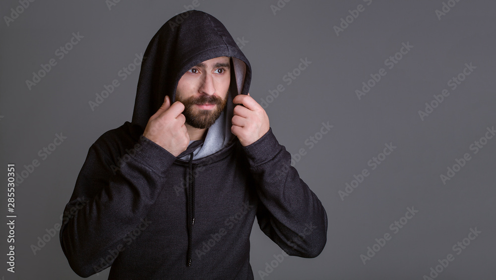 One man in a black sweater on a gray background. Hands puts on a hood, a face with a beard. One man in a black sweater on a gray background. Hands puts on a hood, a face with a beard.