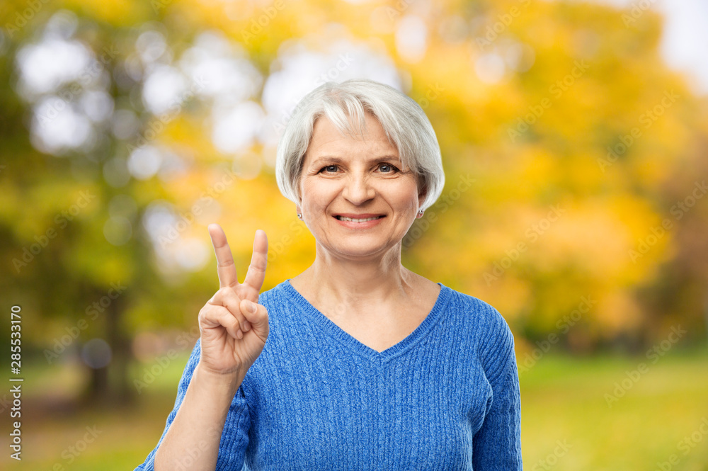 gesture and old people concept - portrait of smiling senior woman in blue sweater showing peace over autumn park background gesture and old people concept - portrait of smiling senior woman in blue sweater showing peace over autumn park background