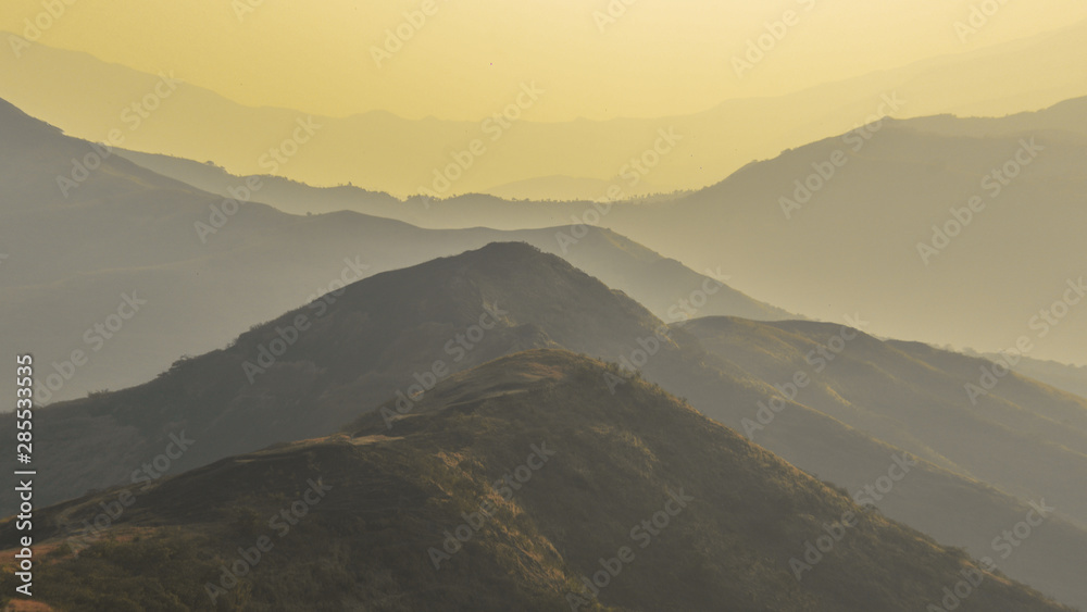 A layer of hills and mountains seen during the evening golden hour during sunset. The hills & mountains can be seen coveredin fog and mist, with a golden yellow sky & the horizon in the background