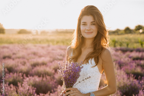 Caucasian Young Woman on Lavender Field Background. Brunette Girl Smiling Looking at Camera Waist Up Shot. Female Model with Natural Makeup Hold Bouquet. Blooming Flower Row Background Bokeh