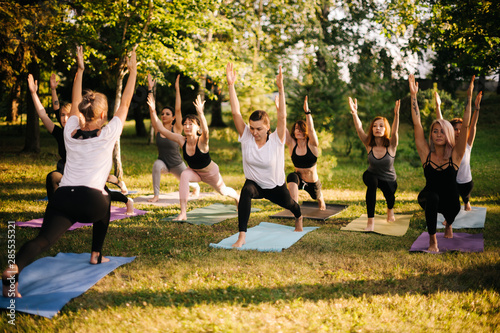 Group of young sporty handsome women practicing yoga lesson with instructor in city park on summer sunny morning. Group of people are standing together in Virabhadrasana 1 exercise, Warrior one pose photo