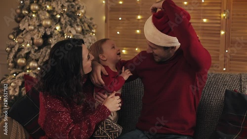 Cheerful Family of Mum Dad and Adorable Baby Boy Sitting on the Sofa near Christmas Tree at Cosy Home Holiday Background. Dad Playes with Son Using his Hat Concept of Holidays and New Year. photo