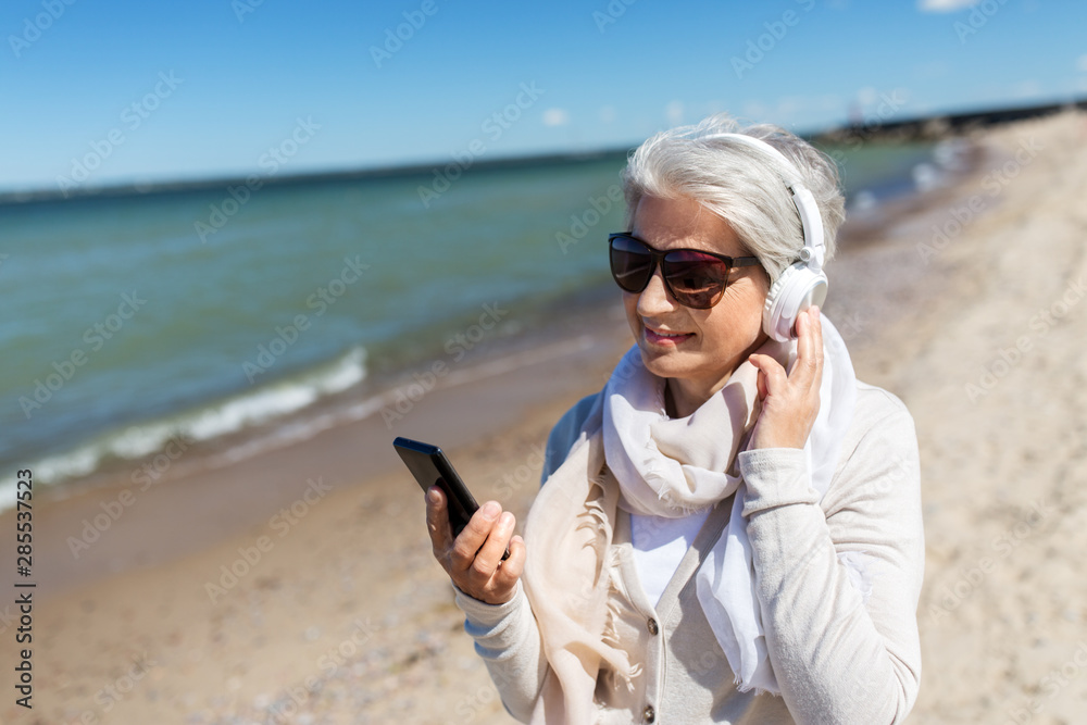 technology, old people and leisure concept - senior woman in headphones and sunglasses listening to music on smartphone on summer beach in estonia