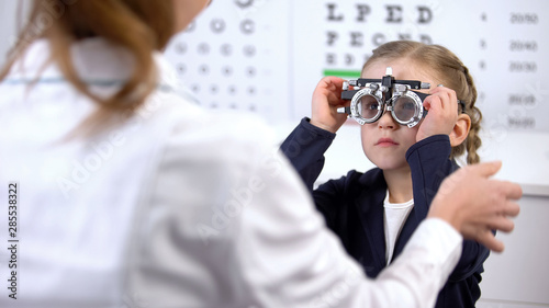 Pretty little schoolgirl visiting oculist for measuring eyesight with phoropter