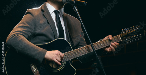 Man with a beard in a jacket plays and sings while standing on stage on a 12-string acoustic guitar. Dark background photo