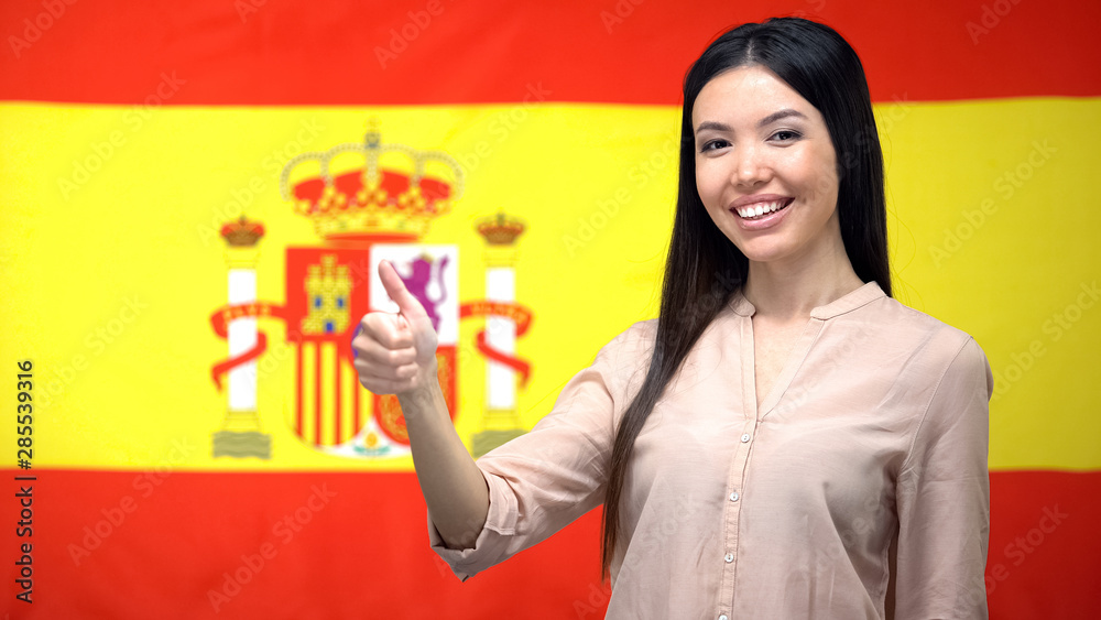 Smiling Asian woman showing thumbs-up gesture against Spanish flag background Smiling Asian woman showing thumbs-up gesture against Spanish flag background