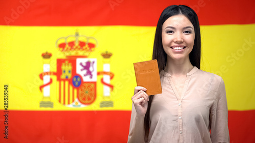 Fotografie Smiling Asian girl holding passport against Spanish flag background, citizenship