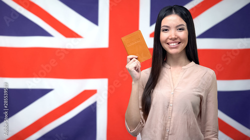 Smiling Asian girl holding passport against British flag background, citizenship
