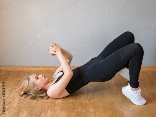 Slender charming young blonde women in black tracksuit and white sneakers posing in front of camera in studio with dumbbells. girl is training at home.