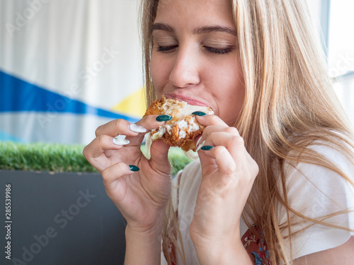 Slender charming young blonde in a white T-shirt and multi-colored sundress poses in front of camera in fast food cafe. girl eating hamburger one by one
