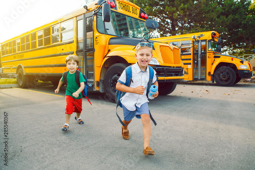 Two funny happy Caucasian boys students kids running near yellow bus on 1 September day. Education and back to school concept. Children friends pupils ready to learn and study.
