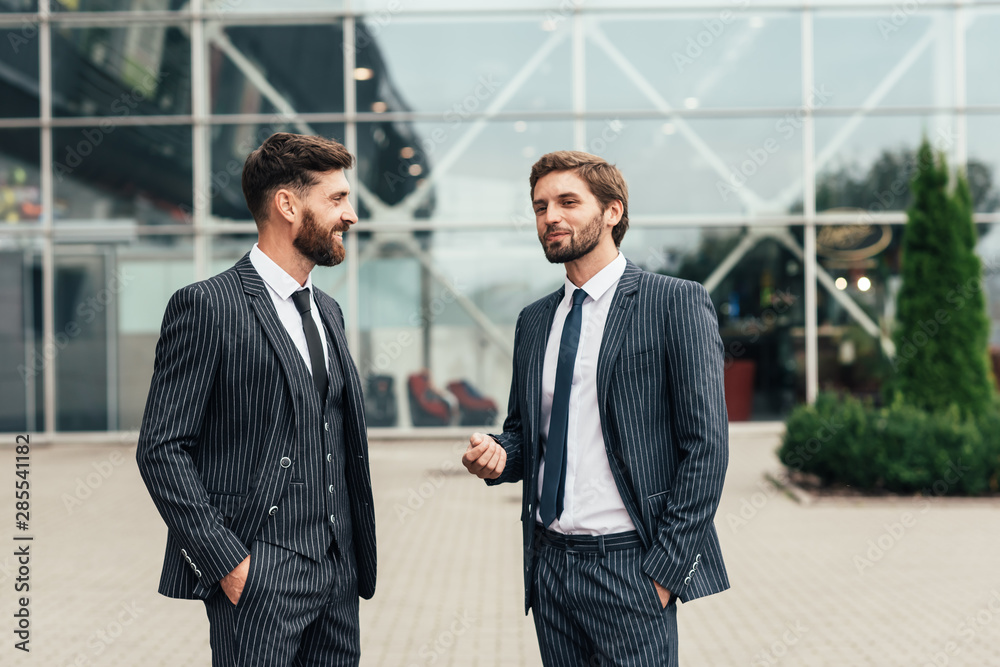 Two smiling businessman colleagues in a blue suit talking and walking business people discussing strategy in a modern city Two smiling businessman colleagues in a blue suit talking and walking business people discussing strategy in a modern city