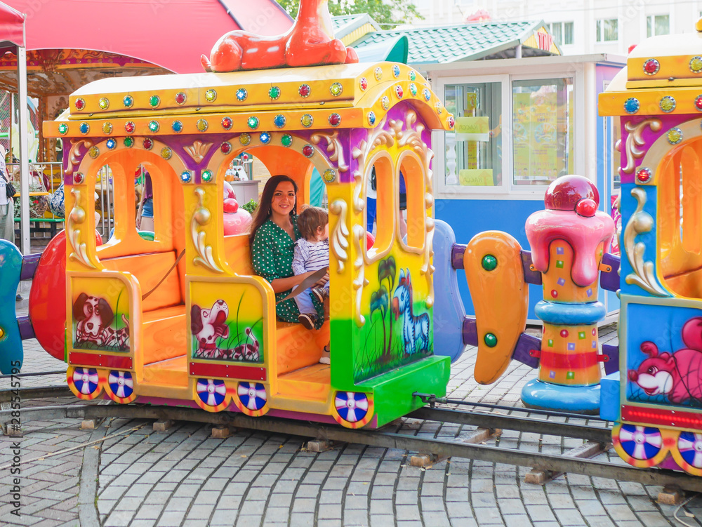 Mom rides on a children's train with a child. Mom rides with a child on a carousel. Portrait of a happy mother and son riding on a carousel Mom rides on a children's train with a child. Mom rides with a child on a carousel. Portrait of a happy mother and son riding on a carousel