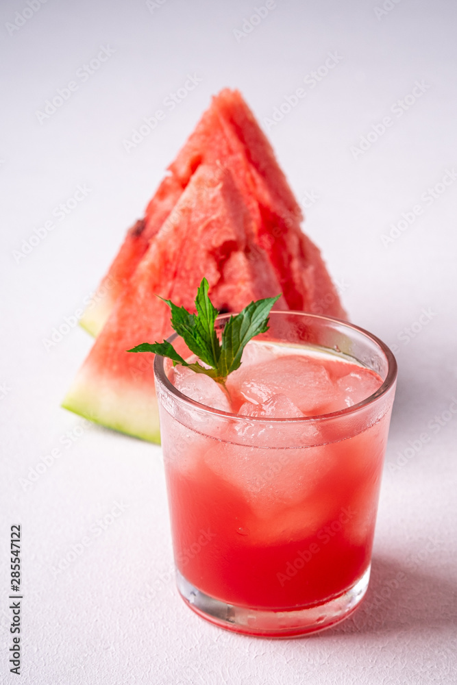 Fresh cold watermelon juice with ice cubes and green mint leaf in glass drink close to two slices of watermelon on white background, copy space, angle view, selective focus Fresh cold watermelon juice with ice cubes and green mint leaf in glass drink close to two slices of watermelon on white background, copy space, angle view, selective focus