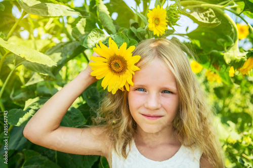 Beautiful happy blonde little girl on sunflower field. Girl holds flower in hand. 