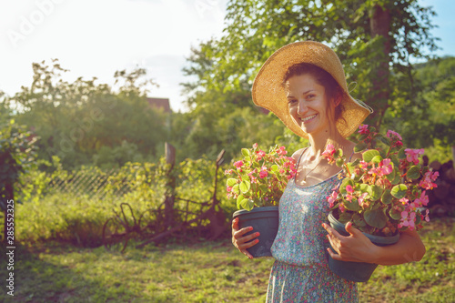 Young woman female farmer happy smiling carry two pots with flowers by her house yard garden flower pot taking care of the plants wearing hat and summer dress