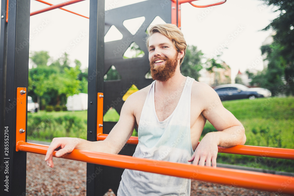 Portrait young bearded man standing on public sports ground and training on parallel bars. Handsome workout athlete. Portrait sporty man with beard, rest after training. Summer workout outdoors