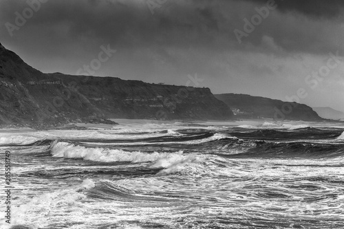 A huge ocean waves breaking on the coastal cliffs in at the cloudy stormy day. Breathtaking romantic seascape of ocean coastline. Bw photo.