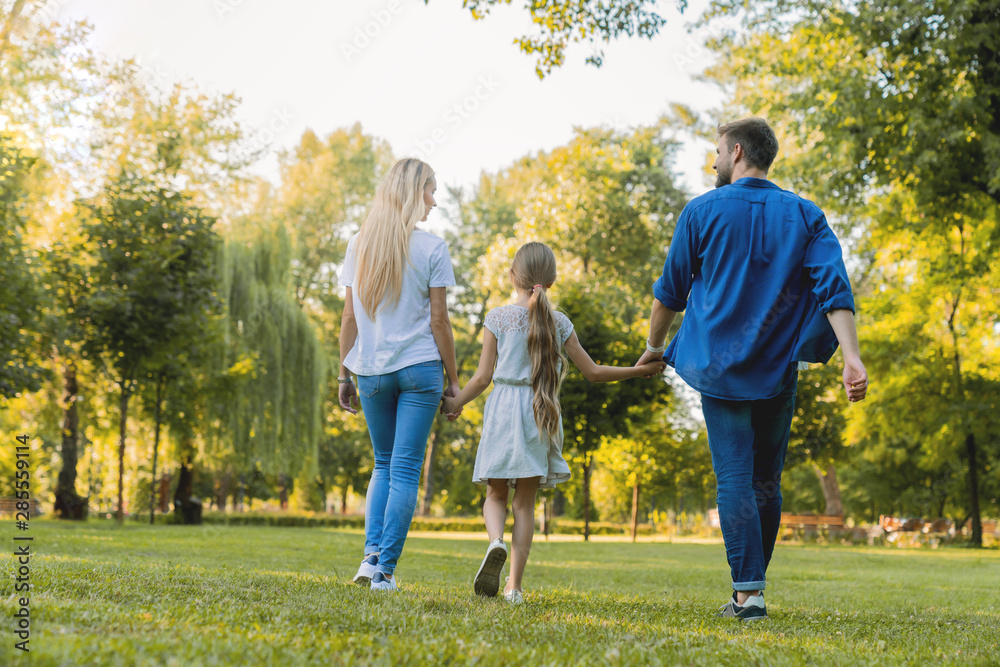 Fototapeta premium Rear view of parents and cute daughter having fun and walking at city park