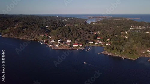 Aerial, drone shot, tilting towards a boat driving on the sea, in front of a small harbor and red buildings, in Ronnes, near Grimstad city, on a sunny day, in Aust Agder, Norway photo