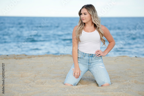 Stunning beautiful woman with blonde hair poses on beach in white tank top and blue jeans - kneeling in sand