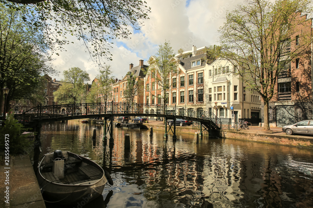 Fototapeta premium Sunday morning in Amsterdam, view of the boat moored at the canal, bridge and townhouses in the background, cloudy sky, warm colours
