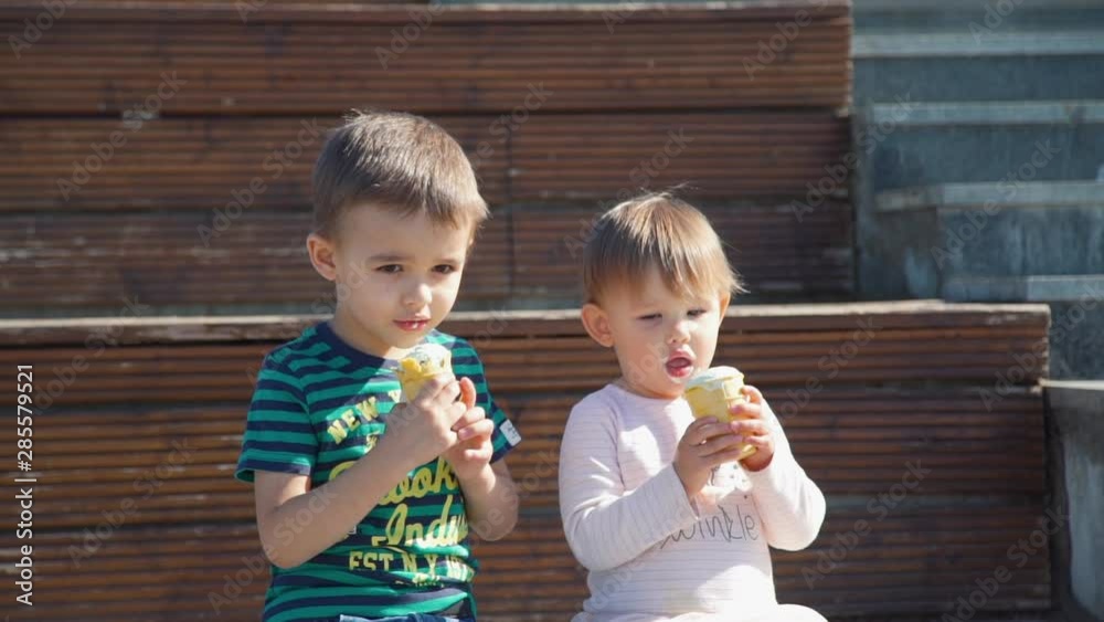 Brother and Sister Eating Ice Cream on the Street while Sitting on the Bench in Slow Motion. Happy Childhood concept Brother and Sister Eating Ice Cream on the Street while Sitting on the Bench in Slow Motion. Happy Childhood concept