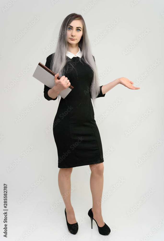 Full length studio portrait of a pretty manager girl with long beautiful hair in a black dress with a folder in her hands on a white background. He stands in front of the camera, smiles, shows hands. Full length studio portrait of a pretty manager girl with long beautiful hair in a black dress with a folder in her hands on a white background. He stands in front of the camera, smiles, shows hands.