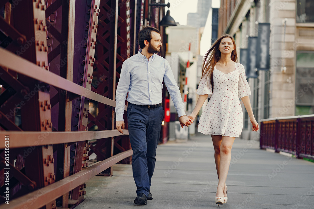beautiful long-haired girl in summer dress with her handsome husband in white shirt and pants walking in sunny Chicago near the big bridge