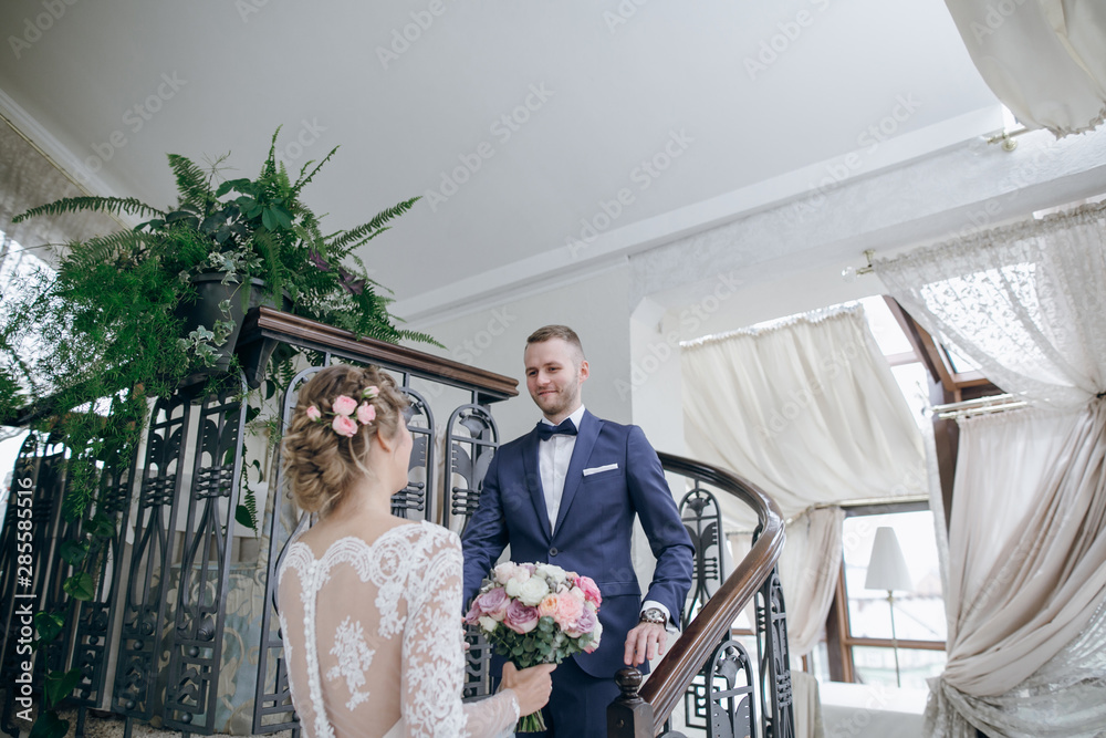Fototapeta premium beautiful and elegant blonde bride in a long white dress with her handsome bride in a blue suit standing in the hotel room with bouquet of flowers