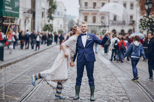 beautiful and elegant blonde bride in a long white dress with her handsome bride in a blue suit wallkig in a raining city with umbrella