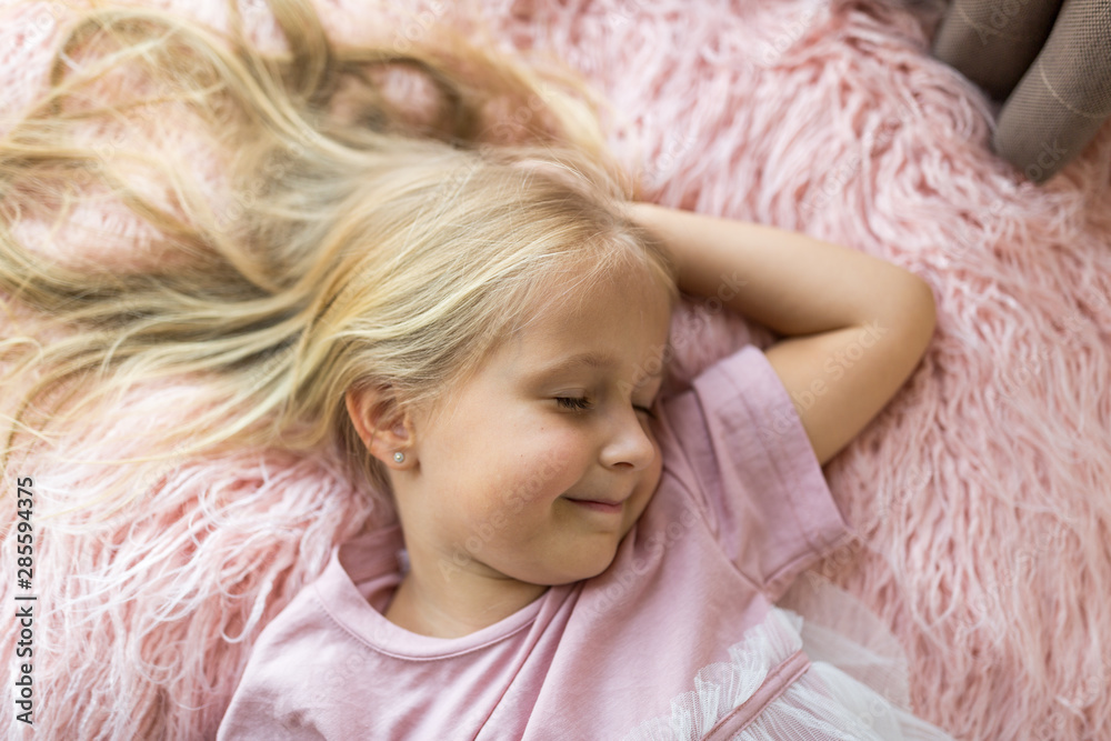 Cute little girl with long blonde hair lying on pink fur blanket with closed eyes pretending sleeping. Happy childhood , children day concept. time for rest, relax, dream, making wishes