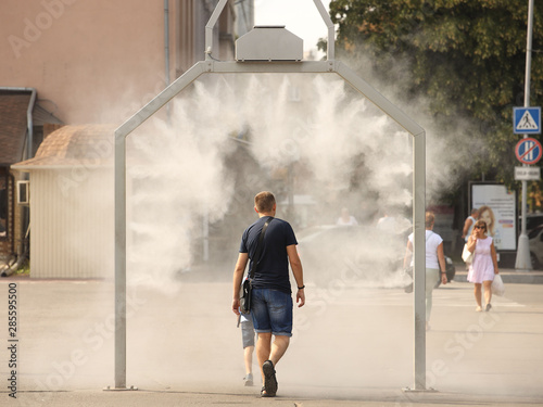 A man in summer clothes goes through the frame of a refreshing street shower. Use fine water dust during the hot season to refresh pedestrians. Protection against overheating and sunstroke by splashin
