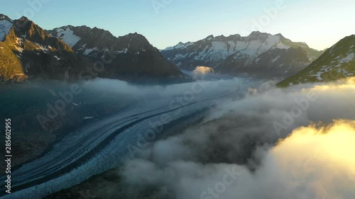 Aerial, reverse, drone shot, of the Aletsch glacier, surrounded by snowy mountain tops and fog clouds, at sunset, on a sunny, summer evening, in the Swiss alps of Valais, Switzerland photo