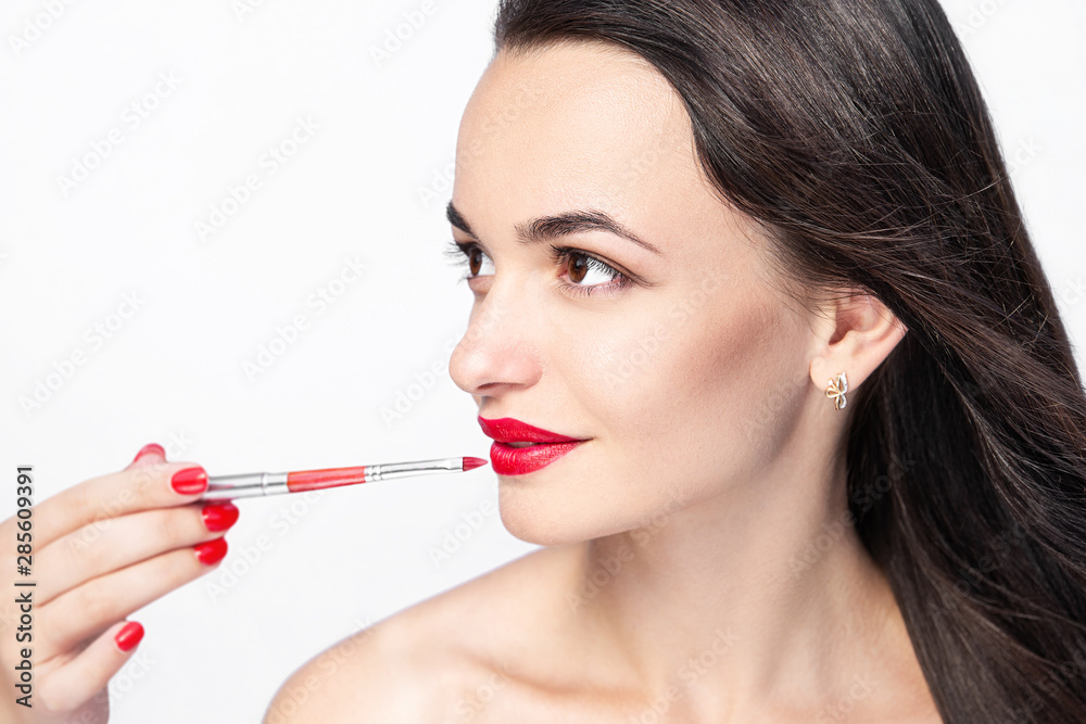 Fototapeta premium Makeup artist does makeup to a beautiful young woman. Brush red lipstick. Female beauty portrait on the white background